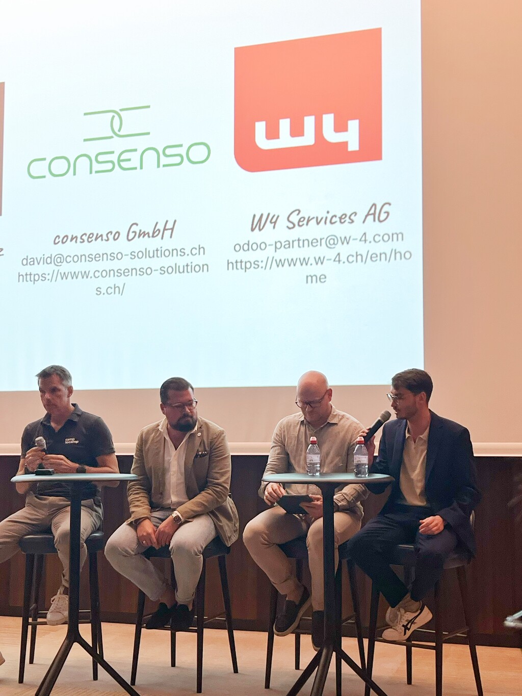 Vier Männer sitzen auf Barhockern an einem Tisch und nehmen an einer Podiumsdiskussion oder einer Q&A-Runde teil. Im Hintergrund werden die Logos und Kontaktinformationen der Unternehmen 