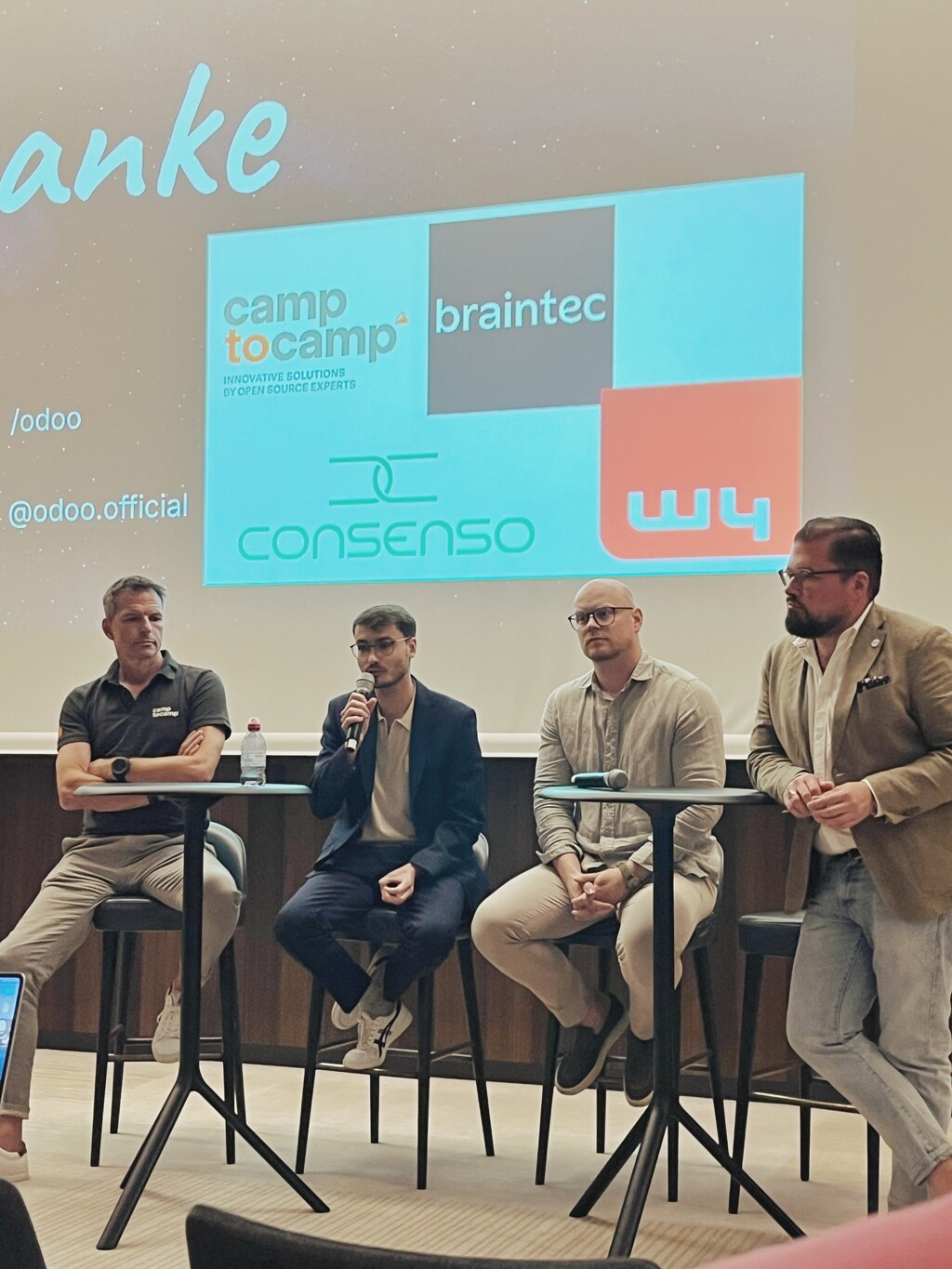 Vier Männer auf einer Bühne, die an einer Podiumsdiskussion teilnehmen. Ein Mann spricht in ein Mikrofon. Im Hintergrund werden auf einer Leinwand die Logos der Unternehmen 