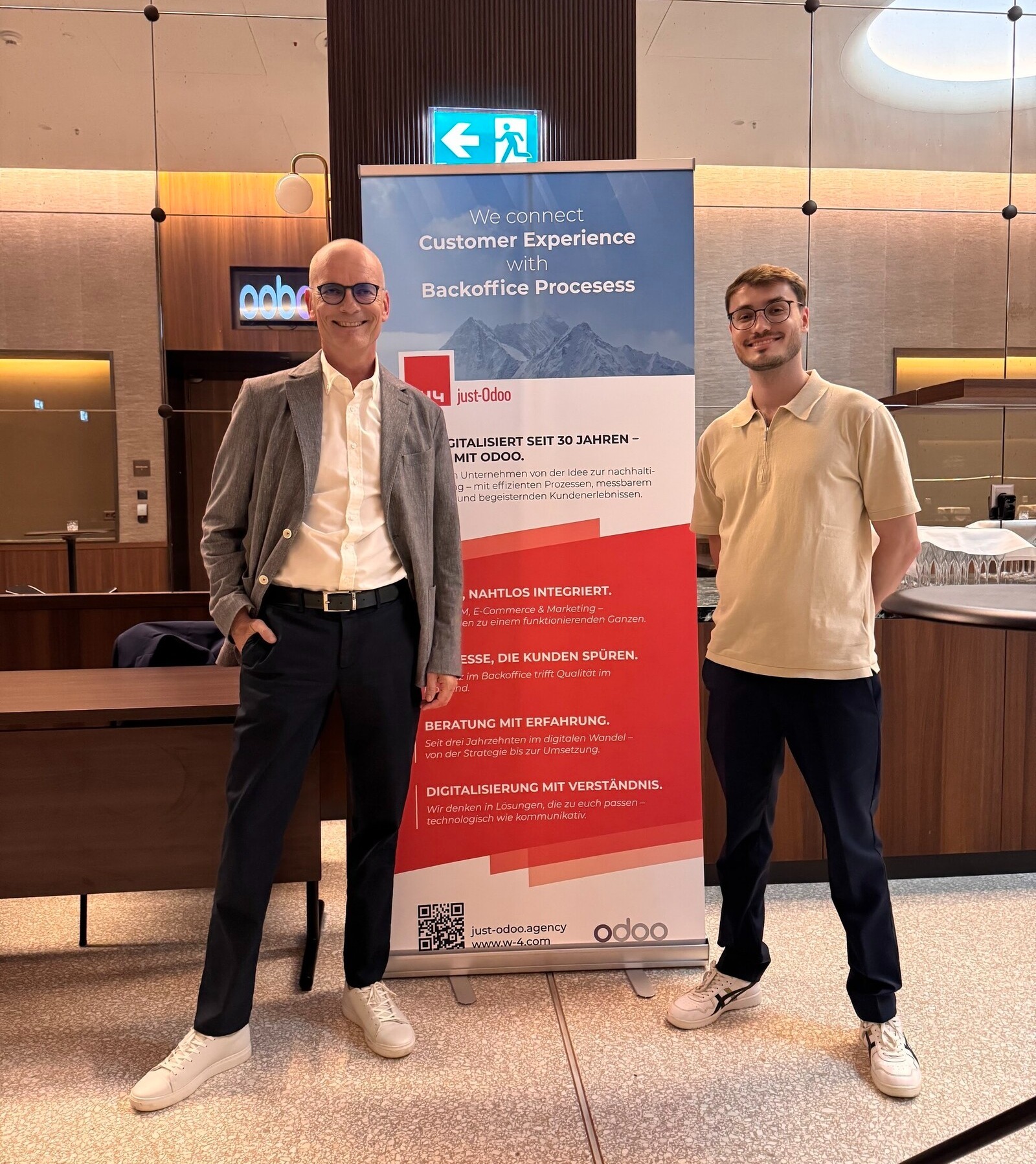 Jörg Wenzel und Ares Wenzel stehen vor einem Roll-up-Banner, das für Odoo-Dienstleistungen wirbt. Das Banner trägt den Slogan „We connect Customer Experience with Backoffice Processes“ und Listeneinträge auf Deutsch zur Digitalisierung und Beratung.